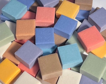 May include: A collection of small, square mosaic tiles in various pastel colors, including blue, pink, yellow, and green. The tiles are arranged in a pile, showcasing their smooth, matte surfaces. These tiles are suitable for craft projects.