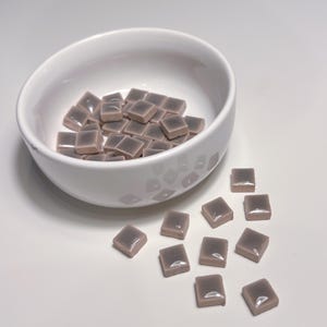 May include: A white ceramic bowl containing small, square, mauve-coloured tiles. Some tiles are scattered outside the bowl. The tiles have a glossy finish, and the bowl has a simple, modern design.