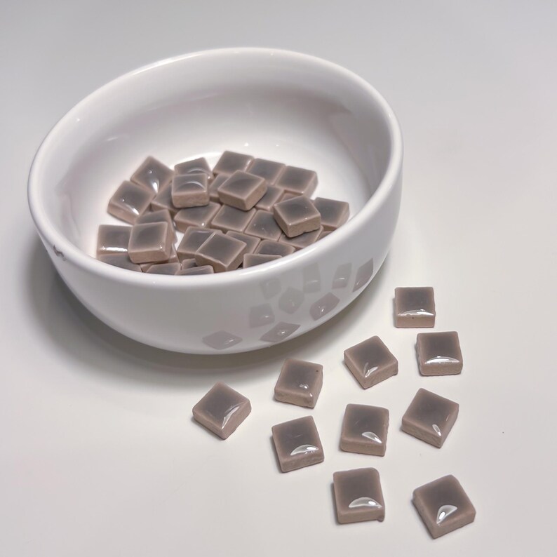 May include: A white ceramic bowl containing small, square, mauve-coloured tiles. Some tiles are scattered outside the bowl. The tiles have a glossy finish, and the bowl has a simple, modern design.