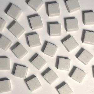 May include: An overhead shot shows numerous small, square, off-white tiles scattered across a white surface. The tiles are uniform in size and shape, with slightly rounded edges. The lighting is bright, highlighting the texture of the tiles.