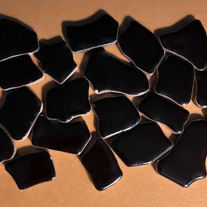 May include: A collection of irregularly shaped, glossy black tiles with silver edges. The tiles are scattered across a neutral-toned surface, creating a mosaic or decorative element. The shapes vary, adding visual interest to the composition.