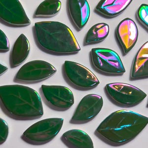 May include: A collection of green, leaf-shaped decorative tiles with iridescent surfaces. The tiles vary in size and shape, with visible veining details. The glossy finish reflects a spectrum of colors, including pink, purple, and yellow, against a white background.