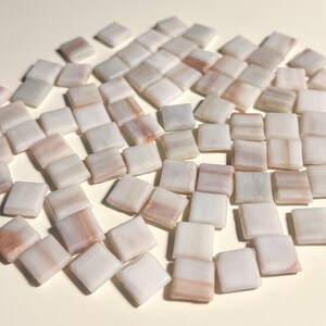 May include: A collection of small, white, square glass tiles with subtle pink and brown streaks. The tiles are scattered on a white surface.