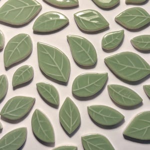 May include: A collection of light green ceramic leaf tiles with white veining details. The leaves vary in size and are arranged on a white surface. These decorative tiles are suitable for various craft and home decor projects.