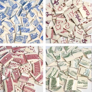 May include: A collection of broken ceramic tiles with various designs. Some tiles feature blue floral patterns, others have red star and stripe motifs, and some display green botanical illustrations. Several tiles have handwritten text in red.