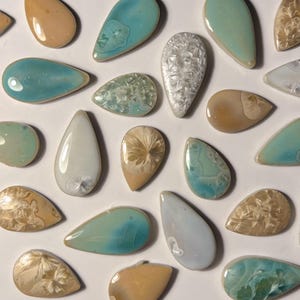 Può includere: Un assortimento di ciondoli in ceramica a forma di goccia in vari colori, tra cui azzurro, beige e bianco. Alcuni ciondoli hanno superfici strutturate o decorate, mentre altri sono lisci e lucidi. I ciondoli sono disposti su una superficie bianca.