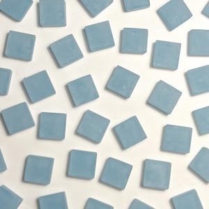 Puede incluir: Una colección de baldosas cuadradas de color azul claro dispersas sobre una superficie blanca. Las baldosas están espaciadas uniformemente y tienen un acabado mate. La imagen es una vista superior, que muestra el patrón geométrico creado por las baldosas.