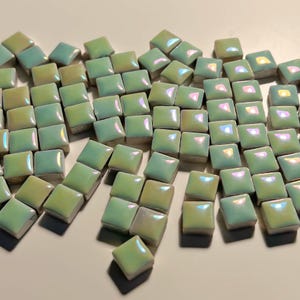 May include: A collection of small, square, iridescent green ceramic tiles. The tiles are scattered on a white surface.