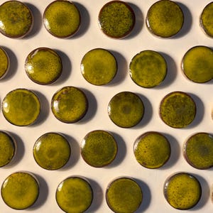 May include: Close-up of multiple olive-green, round ceramic tiles. Each tile has a glossy finish and a dark brown edge. The tiles are arranged in a grid pattern, creating a visually appealing and textured surface. The tiles are likely for a mosaic or decorative project.