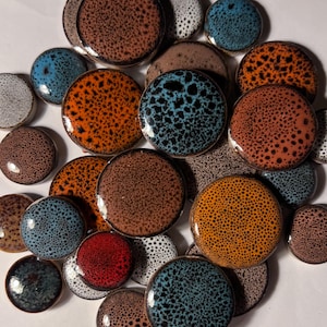 Puede incluir: Una colección de pomos redondos de cerámica en varios colores, incluyendo naranja, marrón, azul y blanco. Cada pomo presenta un patrón moteado y un acabado brillante. Los pomos están dispuestos en un primer plano, mostrando sus diversos tonos y texturas.