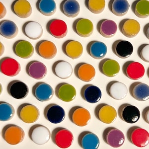 Puede incluir: Una colección de botones pequeños, circulares y de parte superior plana dispuestos en un patrón de cuadrícula. Cada botón presenta una superficie lisa, brillante y de color sólido en varios tonos, incluyendo rojo, azul, amarillo, verde, naranja, morado, blanco y negro.