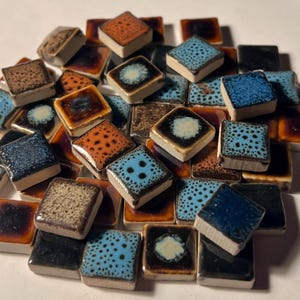 May include: A collection of small, square ceramic tiles in various colors and patterns. The tiles feature glossy finishes in shades of brown, orange, blue, and black. Some tiles have speckled or dotted designs, while others have a central circular pattern.