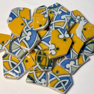 Könnte beinhalten: Eine Ansammlung zerbrochener Keramikfliesen mit einem dekorativen Muster. Die Fliesen weisen eine lebendige Farbpalette aus Gelb, Blau und Weiß auf. Das Design umfasst geometrische Formen und florale Elemente, wodurch ein Mosaikeffekt entsteht.