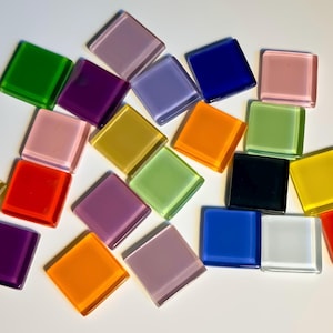 May include: Assortment of colorful square glass tiles in various shades of pink, purple, blue, green, yellow, orange, red, and black. The tiles are arranged in a random pattern on a white surface.