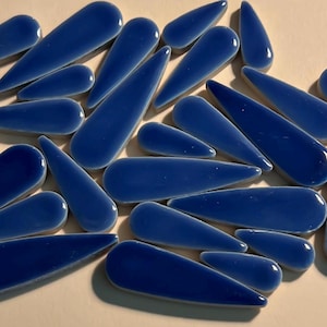 May include: A collection of glossy, teardrop-shaped, cobalt blue tiles. The tiles vary in size and are scattered across a neutral surface. The smooth, curved shapes and vibrant color suggest they are decorative elements for mosaic art or crafts.