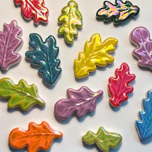 Op de afbeelding: Een verzameling kleurrijke, iriserende keramische eikenbladvormen. De bladeren zijn in verschillende kleuren, waaronder rood, geel, groen, blauw en paars. Elk blad heeft een glanzende afwerking en ligt op een witte ondergrond.