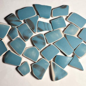 Könnte beinhalten: Eine Sammlung unregelmäßig geformter, teal-farbener Keramikteile mit dunkelgrauen, wirbelartigen Mustern. Jedes Teil hat einen weißen Rand, wodurch ein mosaikartiges Aussehen entsteht. Die Teile sind vor einem weißen Hintergrund verstreut.