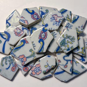 May include: A collection of white ceramic tile shards with floral patterns in pink, blue, and yellow. The broken pieces have a geometric shape and are scattered across a white surface. The design features stylized flowers and leaves.