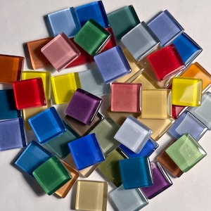 May include: A collection of small, square glass tiles in a variety of translucent and opaque colors, including red, blue, yellow, green, purple, orange, and clear tones, scattered in a random arrangement with smooth, flat surfaces.