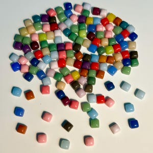May include: A large pile of colorful square glass tiles in various shades of blue, green, red, yellow, brown, pink, and white. The tiles are scattered on a white surface.