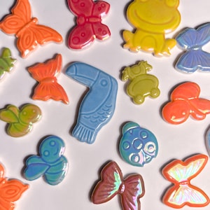 Könnte beinhalten: Eine Sammlung farbenfroher Keramikfiguren von Tieren und Insekten. Das Sortiment umfasst Schmetterlinge, einen Frosch, einen Tukan, einen Marienkäfer und andere Kreaturen in Orange, Blau, Gelb und Grün.