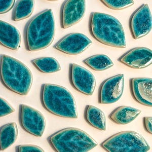 May include: Turquoise leaf-shaped ceramic tiles with a glossy finish. The tiles have a textured surface with visible veining, and are arranged on a white background. These decorative tiles are suitable for mosaics and craft projects.