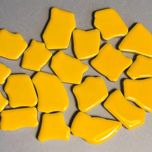 May include: A collection of irregularly shaped, glossy yellow ceramic tiles. The tiles vary in size and form, with smooth, rounded edges and a bright, sunny color. They are scattered on a neutral gray surface, creating a visually appealing arrangement.