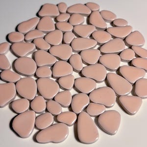 May include: A collection of light pink ceramic tiles in various irregular shapes, resembling pebbles. The tiles are smooth and glossy, with a slightly textured surface.