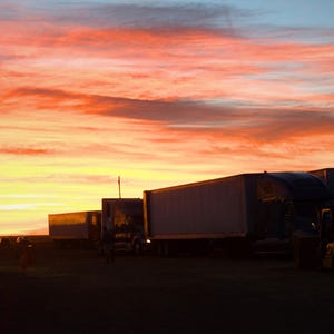 Op de afbeelding: Een levendige zonsondergang boven een truckstop, met een lucht van oranje, gele en blauwe tinten. Silhouetten van vrachtwagens en trailers zijn geparkeerd, met een vorkheftruck zichtbaar. De scène legt een moment van overgang vast.