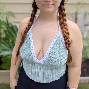 May include: A sage green crochet halter top featuring a deep V-neckline with a white crocheted border trim. The garment is constructed with a textured stitch pattern and includes thin white straps that tie behind the neck for a secure fit.
