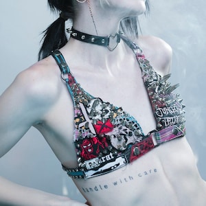 Op de afbeelding: Een zwart-witte punk-bh met een hartvormige choker, bezet met zilveren spikes en versierd met patches. De bh heeft een blauw-wit gestreepte band en de tekst "handle with care" is getatoeëerd op de torso van de drager.