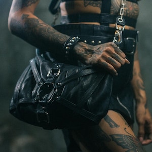 May include: Black leather harness and skirt set with a matching bag. The harness features a chain detail and the bag has a buckle closure. The outfit is worn with a choker and bracelet. The person has tattoos on their arms and legs.