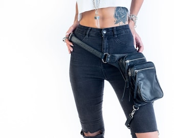 May include: A woman wearing a white tank top with silver chains and a black leather thigh bag. She is wearing black jeans with rips at the knees.