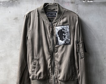 May include: A green bomber jacket with a zipper closure and a black patch with the text "DUFT + BEAU" on the left side of the chest. The jacket has a white and gray image patch on the right side of the chest.
