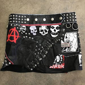 Op de afbeelding: Zwarte leren minirok met zilveren studs, veiligheidsspelden en patches. De patches bevatten een anarchiesymbool, schedels en de tekst "Jungle Tribe" en "Suffolk Tribe".