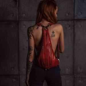 May include: A red leather backpack with multiple zippered compartments. The backpack is worn by a person with tattoos on their back and arms.