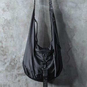 Pode incluir: Uma bolsa preta de ombro com uma alça longa e um fecho de fivela. A bolsa está pendurada numa parede de betão cinzento.