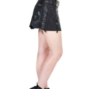 May include: A black leather mini skirt with a decorative design and multiple buckles. The skirt has a high waist and a fitted silhouette. The model is wearing black heeled ankle boots.