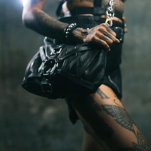 K&ouml;nnte beinhalten: Schwarze Ledertasche mit silberner Hardware. Die Tasche hat eine abgerundete Form mit einem oberen Griff und einem Schultergurt. Die Tasche wird von einer Person mit T&auml;towierungen am Arm gehalten.