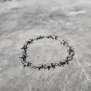 Puede incluir: Un collar de alambre de púas plateado con cierre de broche. El collar está hecho de metal y tiene un aspecto áspero e industrial.