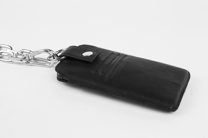 Pu&ograve; includere: Una custodia per telefono in pelle nera con una catena argentata attaccata. La custodia ha una patta con una chiusura a bottone argentata.