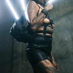 K&ouml;nnte beinhalten: Eine schwarze Lederhandtasche mit runder Form wird &uuml;ber der Schulter getragen. Die Person tr&auml;gt ein schwarzes Lederoutfit, einschlie&szlig;lich Rock, Oberteil und Accessoires. Der Hintergrund ist eine dunkle, strukturierte Wand mit hellen wei&szlig;en Lichtern.