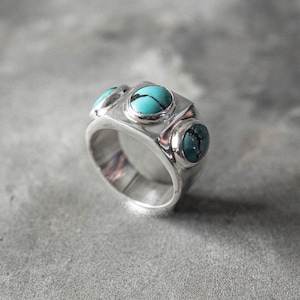 以下が含まれることがあります： 3つのターコイズストーンが付いたシルバーリング。リングは、正方形の中央の石と2つの丸いサイドストーンを備えた幾何学的なデザインです。ターコイズストーンは鮮やかな青緑色です。