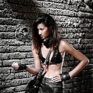 May include: A woman wearing a black leather skirt with a studded design, a black leather halter top, and a black leather bag. She is standing in front of a brick wall.