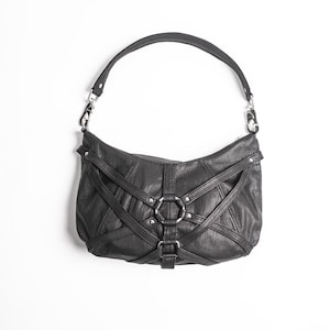 K&ouml;nnte beinhalten: Schwarze Leder-Umh&auml;ngetasche mit einem einzelnen Riemen. Die Tasche hat ein einzigartiges Design mit Riemen und silberfarbenen Metallringen und Beschl&auml;gen. Die Tasche ist vor einem wei&szlig;en Hintergrund platziert.