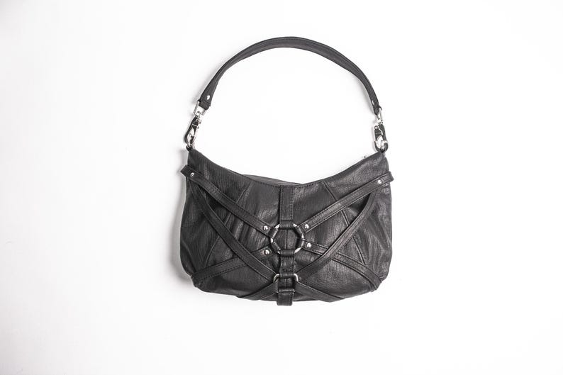K&ouml;nnte beinhalten: Schwarze Leder-Umh&auml;ngetasche mit einem einzelnen Riemen. Die Tasche hat ein einzigartiges Design mit Riemen und silberfarbenen Metallringen und Beschl&auml;gen. Die Tasche ist vor einem wei&szlig;en Hintergrund platziert.