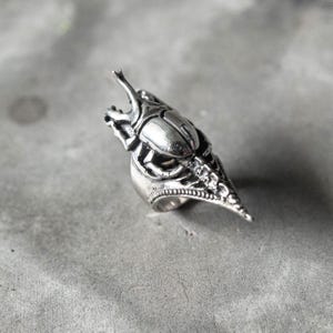 May include: A silver-colored ring with a detailed beetle design. The ring features a pointed tip and intricate insect-like detailing, showcasing a unique and gothic aesthetic. The ring is set against a gray background.
