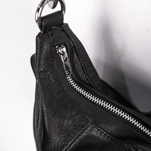 K&ouml;nnte beinhalten: Nahaufnahme einer schwarzen Leder-Umh&auml;ngetasche mit silbernem Rei&szlig;verschluss und Beschl&auml;gen. Die Tasche hat eine strukturierte Oberfl&auml;che und einen silbernen Metallring f&uuml;r den Riemen.