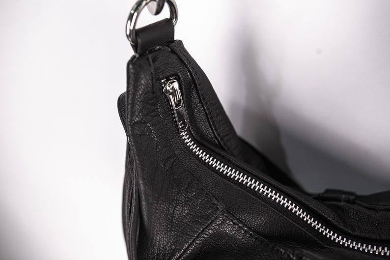 K&ouml;nnte beinhalten: Nahaufnahme einer schwarzen Leder-Umh&auml;ngetasche mit silbernem Rei&szlig;verschluss und Beschl&auml;gen. Die Tasche hat eine strukturierte Oberfl&auml;che und einen silbernen Metallring f&uuml;r den Riemen.
