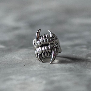 May include: A silver-colored ring with a detailed design of sharp teeth and fangs. The ring has a gothic or fantasy theme, with a focus on the intricate details of the teeth. The ring is likely made of metal.